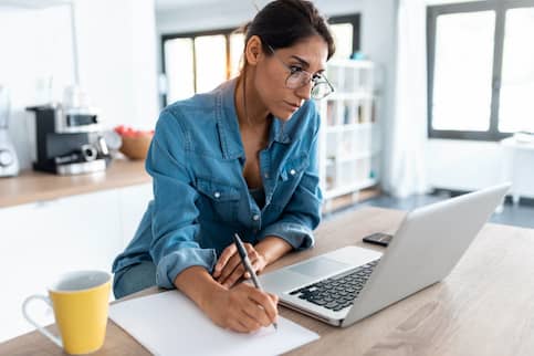 Woman at kitchen table on laptop and writing notes on a piece of paper. 