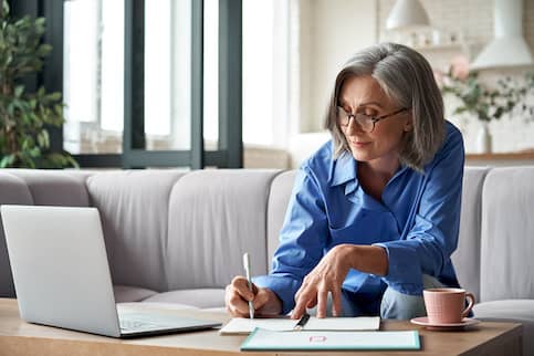 Woman working on a computer sitting on a couch, writing down notes on a coffee table.