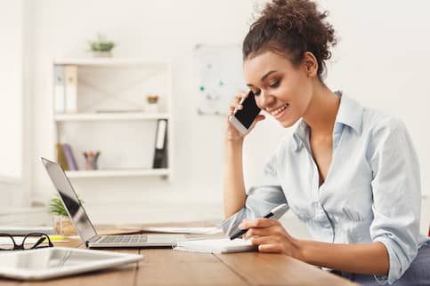 Woman smiling on cellphone while jotting notes down on home office desk.