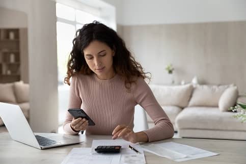 Young woman sitting at table in living room using calculator and laptop computer.