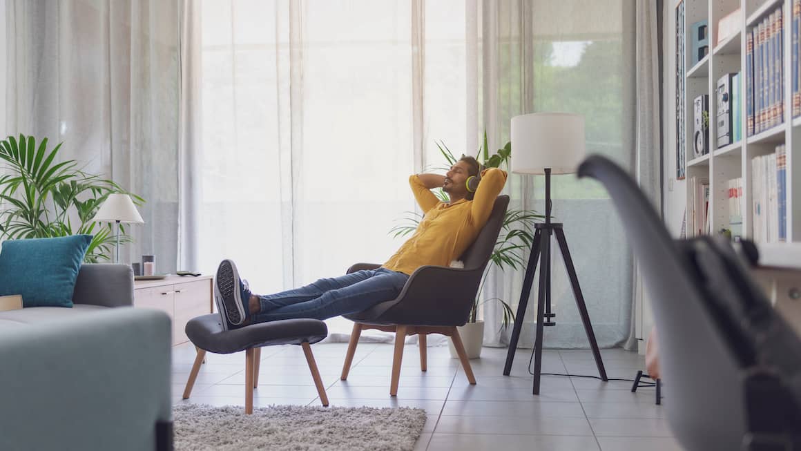 Young adult male lounging in chair in apartment wearing headphones, yellow shirt and jeans.