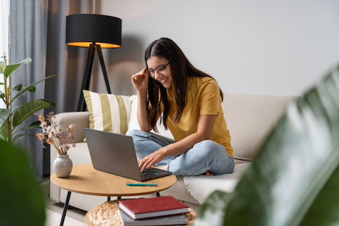 Young woman adjusting her glasses while sitting on couch and working on laptop.
