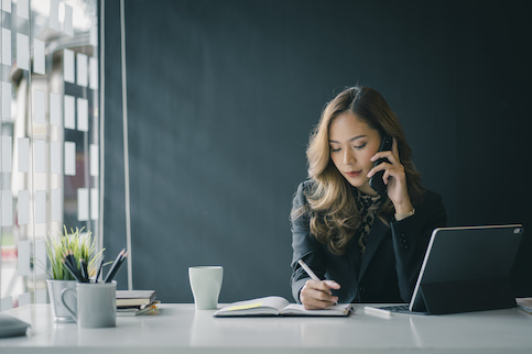 Business woman next to a laptop and talking on her phone while writing in her journal.