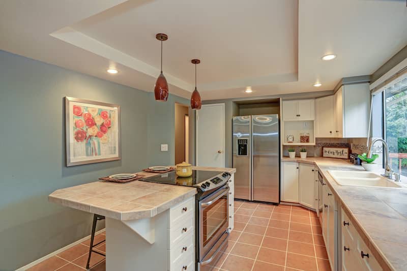 Kitchen with floral painting on blue wall, island with stove, and hanging lights attached to tray ceiling.