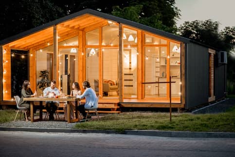 Friends eating outside of home with floor to ceiling windows lit up from inside.