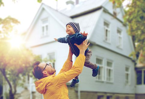 Dad lifting young child into the air in front of large house. 