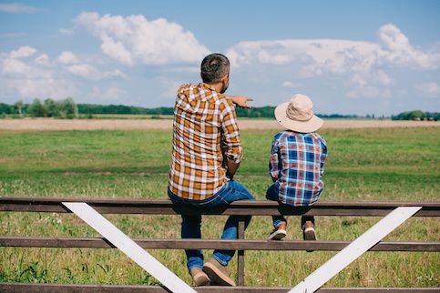 Man and young boy sitting on a fence near a large grassy field.