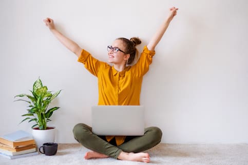 Young woman sitting cross-legged on floor with laptop and arms stretched in air in excitement.