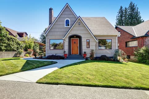 Small tan house with wooden door, tidy landscaping and cement walkway to front door and side of home.