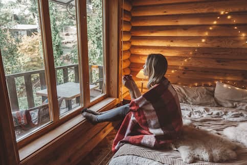 Woman in wooden cabin bedroom looking out of a window and sipping tea.