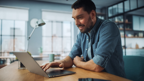 Young man in chambray shirt at kitchen table looking up VA loan refinancing on laptop with smile on face.