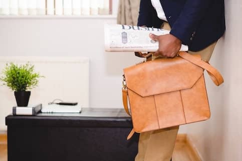 Person holding a tan messenger bag and a rolled up newspaper.