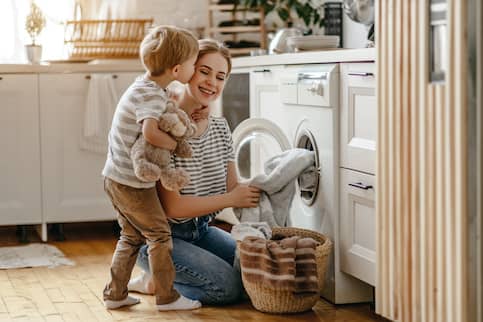 Mom Kneeling At Washing Machine Getting A Kiss By Son With Teddy Bear
