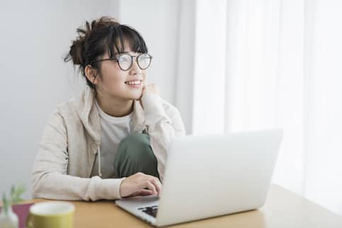 Young Asian Woman With Round Turtle Shell Glasses On Laptop Casually At Table