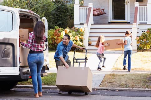 A young couple unloads a moving van and stacks boxes on a hand truck as their two kids carry a large box towards their new home. 