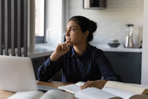 Student at home with laptop and books looking thoughtful.