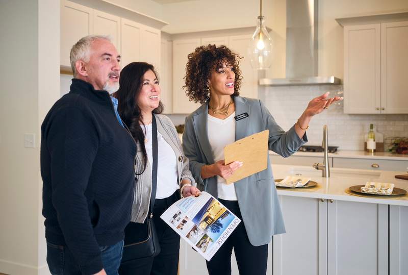 A man and a woman stand in the kitchen of a house for sale, while a real estate agent shows them around.
