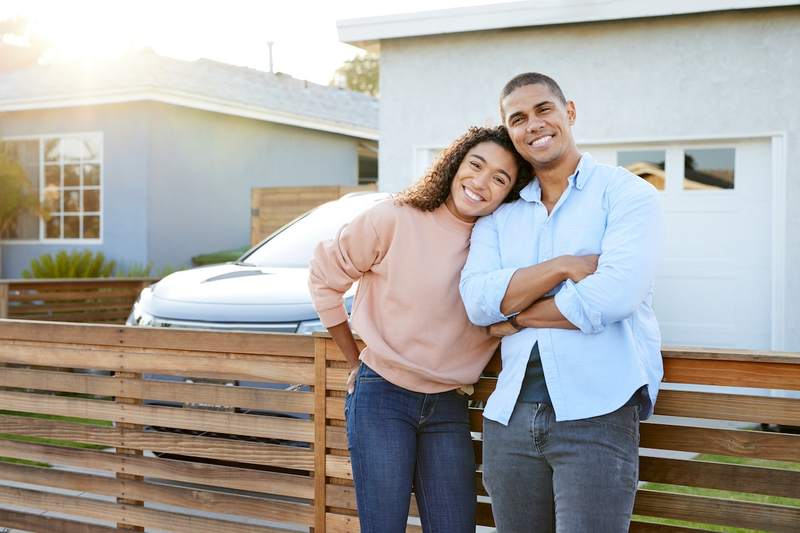 A woman leans her head on the shoulder of a man crossing his arms in front of their car and the fence of their California home.