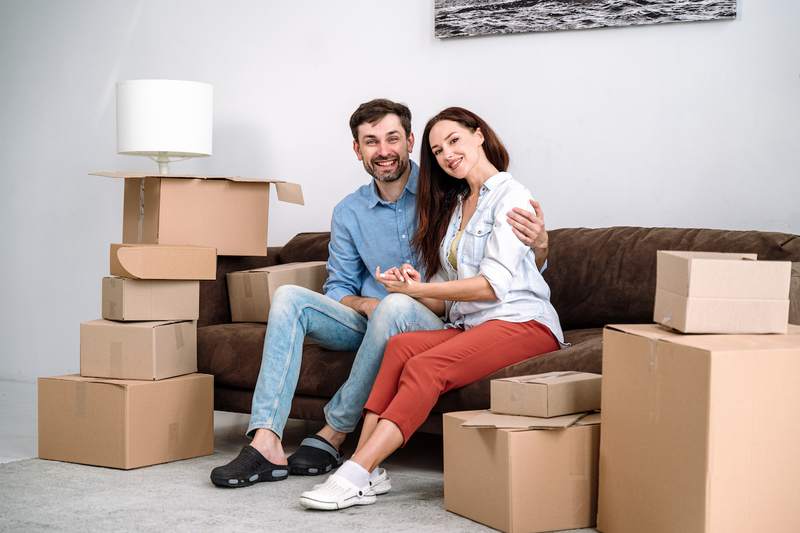 A man and a woman sit together on a sofa, surrounded by moving boxes.