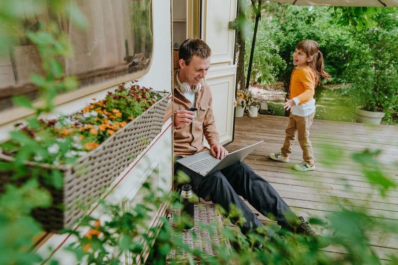 A man with a laptop drinks tea on the patio of a manufactured home with his daughter.