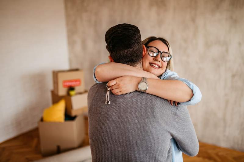 A woman facing the camera hugs her partner, holding the keys to their new home. Moving boxes sit in the corner of the room.