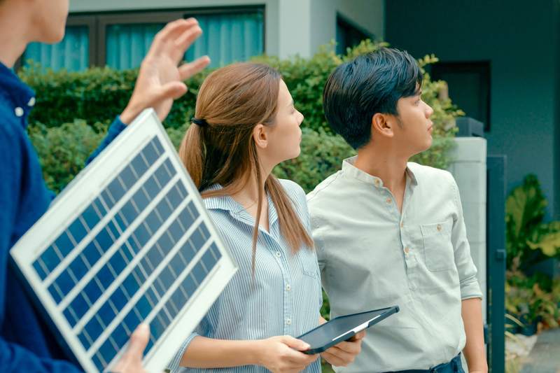 A real estate agent discusses solar panels with a couple looking at the roof of a home.